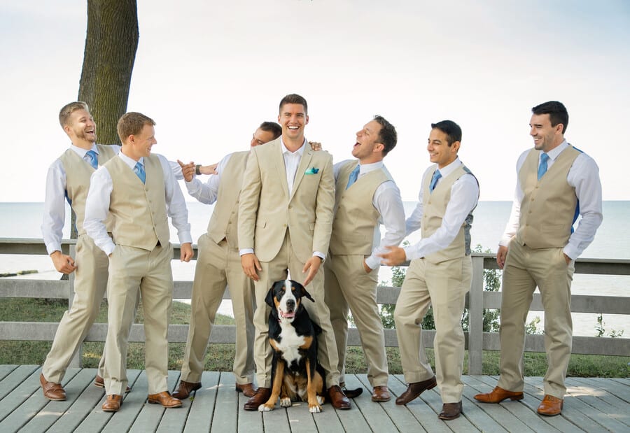 Groomsmen in tan waistcoats and light blue ties laughing on a lakeside boardwalk with a dog sitting in front of the groom