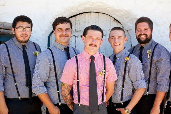 Groom in a pink gingham shirt with a black tie beside groomsmen in blue gingham shirts and black suspenders