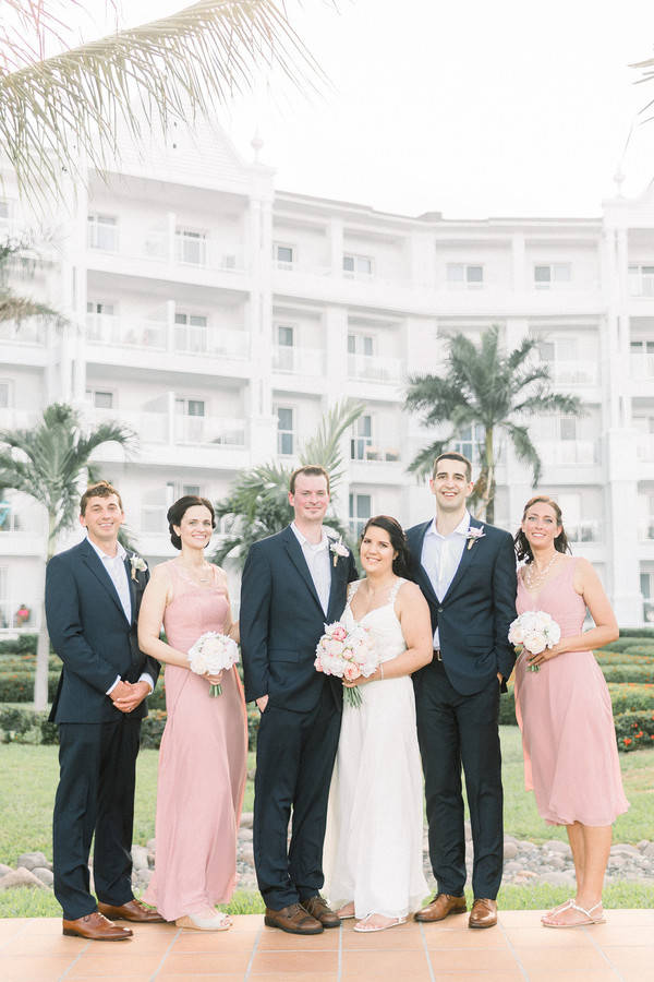 Small wedding party with groom and two groomsmen in navy suits with open collars beside bridesmaids in dusty pink