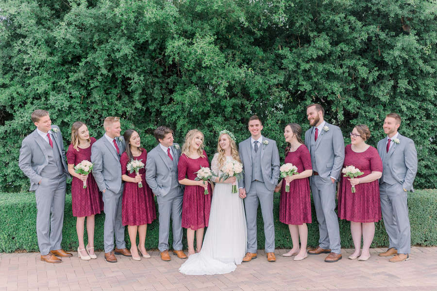 Wedding party alternating groomsmen in light grey three-piece suits with cranberry ties and bridesmaids in cranberry lace dresses