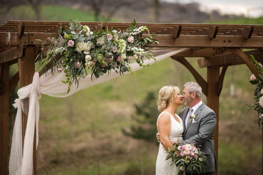 An older bride and groom kissing beneath a wooden arch draped with roses and greenery