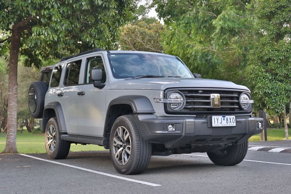 A grey 2026 GWM Tank 300 PHEV SUV with a rear-mounted spare wheel, parked in a car park surrounded by trees.