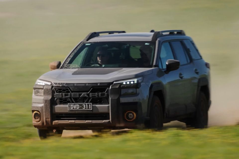 A Subaru Outback Wilderness SUV driving across a grassy field, seen from the front-side, kicking up dust