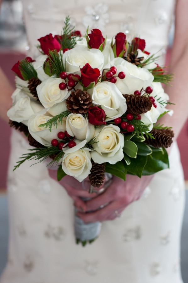 Bridal bouquet of white and red roses with pinecones holly berries and evergreen against a beaded ivory gown