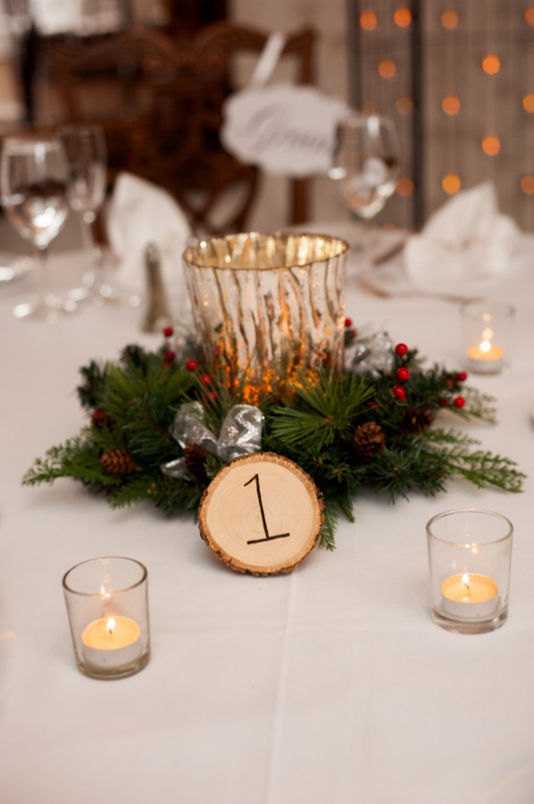 Gold mercury-glass hurricane glowing inside an evergreen wreath with pinecones red berries and a wood-slice table number surrounded by votives