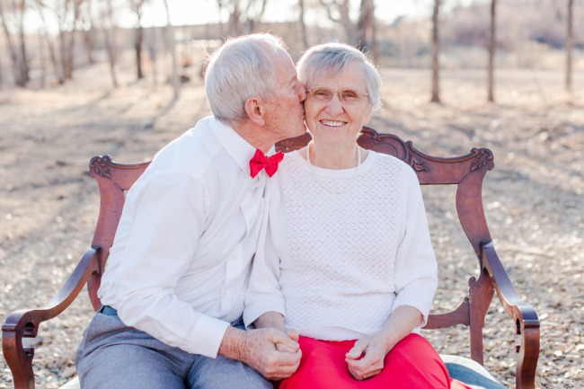 An elderly man kissing his wife's cheek as they sit close together on a bench outdoors, she smiles widely