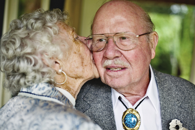 An older couple sharing a cheek kiss, the woman in a patterned blue top kissing the man who wears a suit jacket