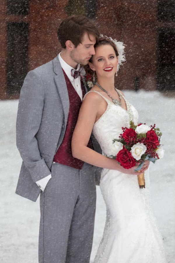 Bride in beaded lace gown with rhinestone statement necklace feather hairpiece and red lipstick holding a red and white rose bouquet beside groom in gray suit with maroon vest as snow falls