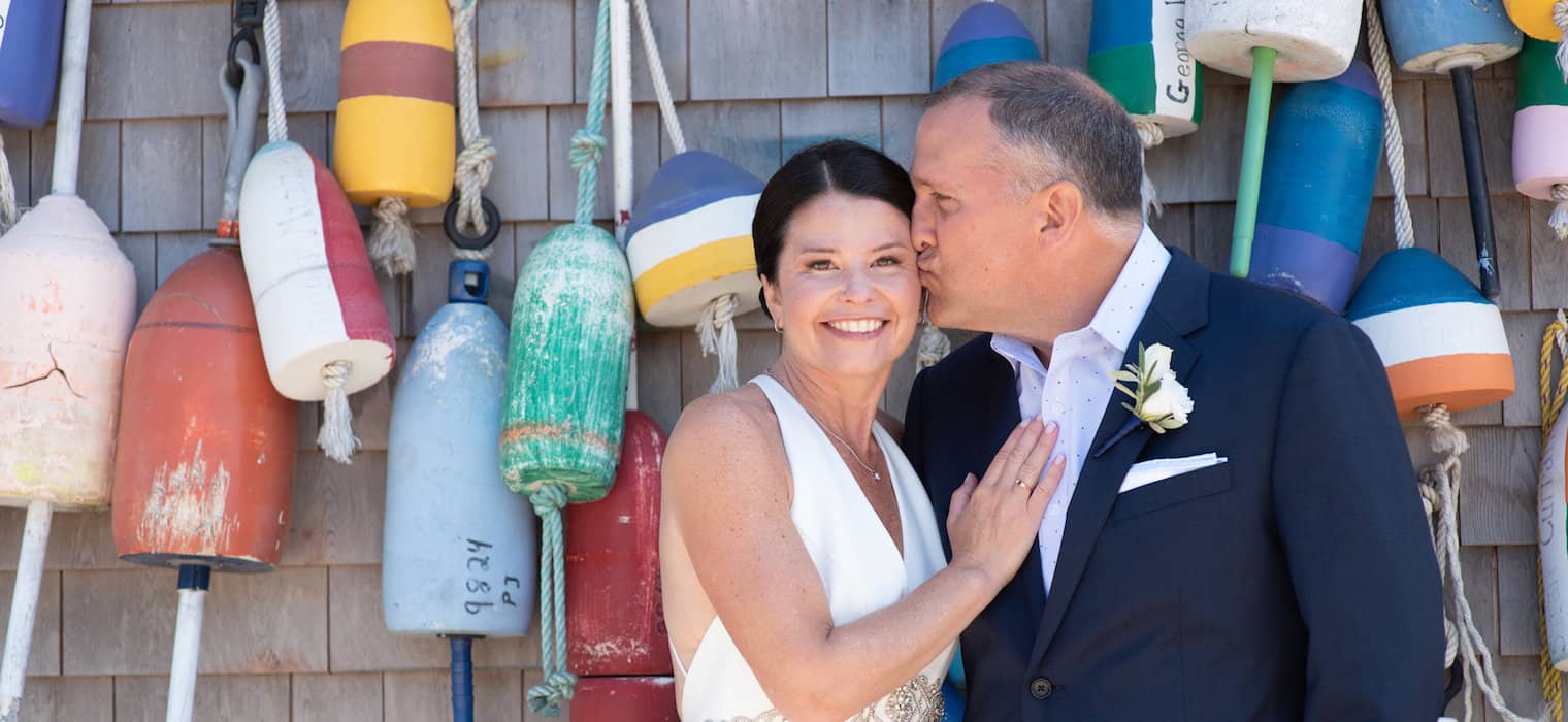 An older bride and groom against a wall of colorful lobster buoys, the groom kissing the bride's cheek as she smiles