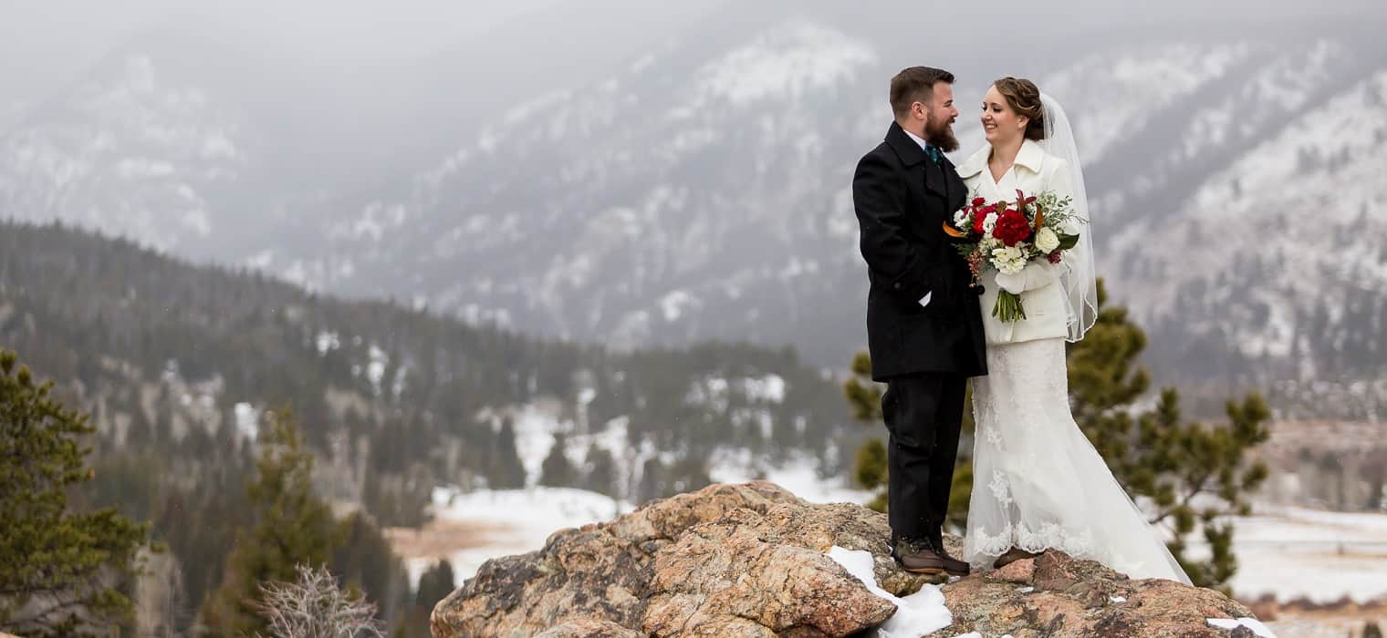 Bride in ivory jacket over lace gown with veil holding a red and white bouquet standing on rocks with snow-dusted mountains behind beside groom in black overcoat