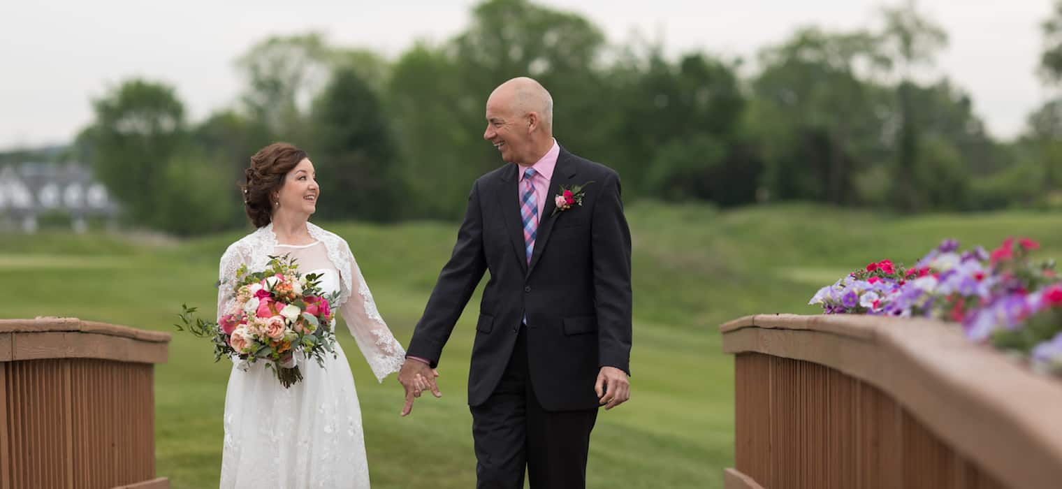 An older bride and groom walking hand in hand across a flower-lined bridge, looking at each other and laughing