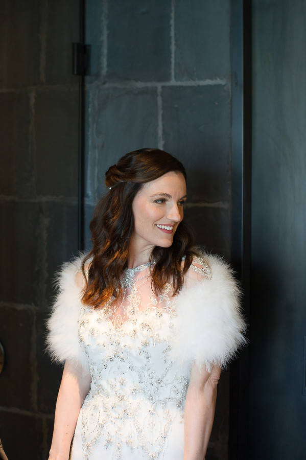 Bride in a white marabou feather shrug over a crystal-beaded illusion gown against a dark slate wall