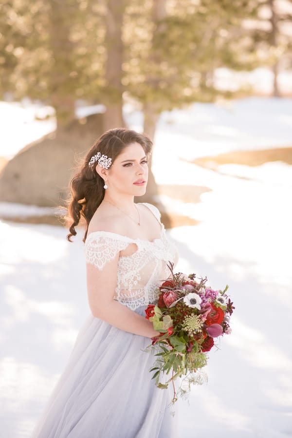 Bride in off-shoulder lace bodice and powder-blue tulle skirt with jeweled hair comb holding a burgundy anemone and protea bouquet in the snow