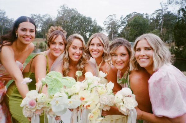 Bride and bridesmaids smiling together outdoors in pastel dresses, holding pale floral bouquets.