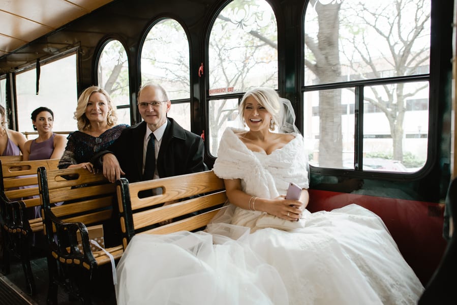 Bride in white fur wrap and tulle ballgown riding a vintage wood-benched trolley with family bare winter trees visible through the arched windows