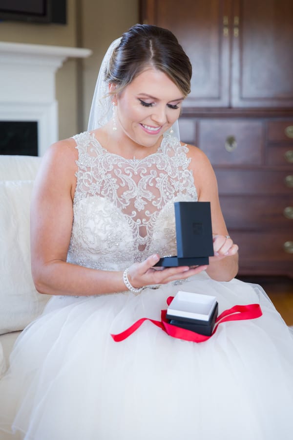 Bride in her gown opening a small black jewelry box with a red ribbon in her lap