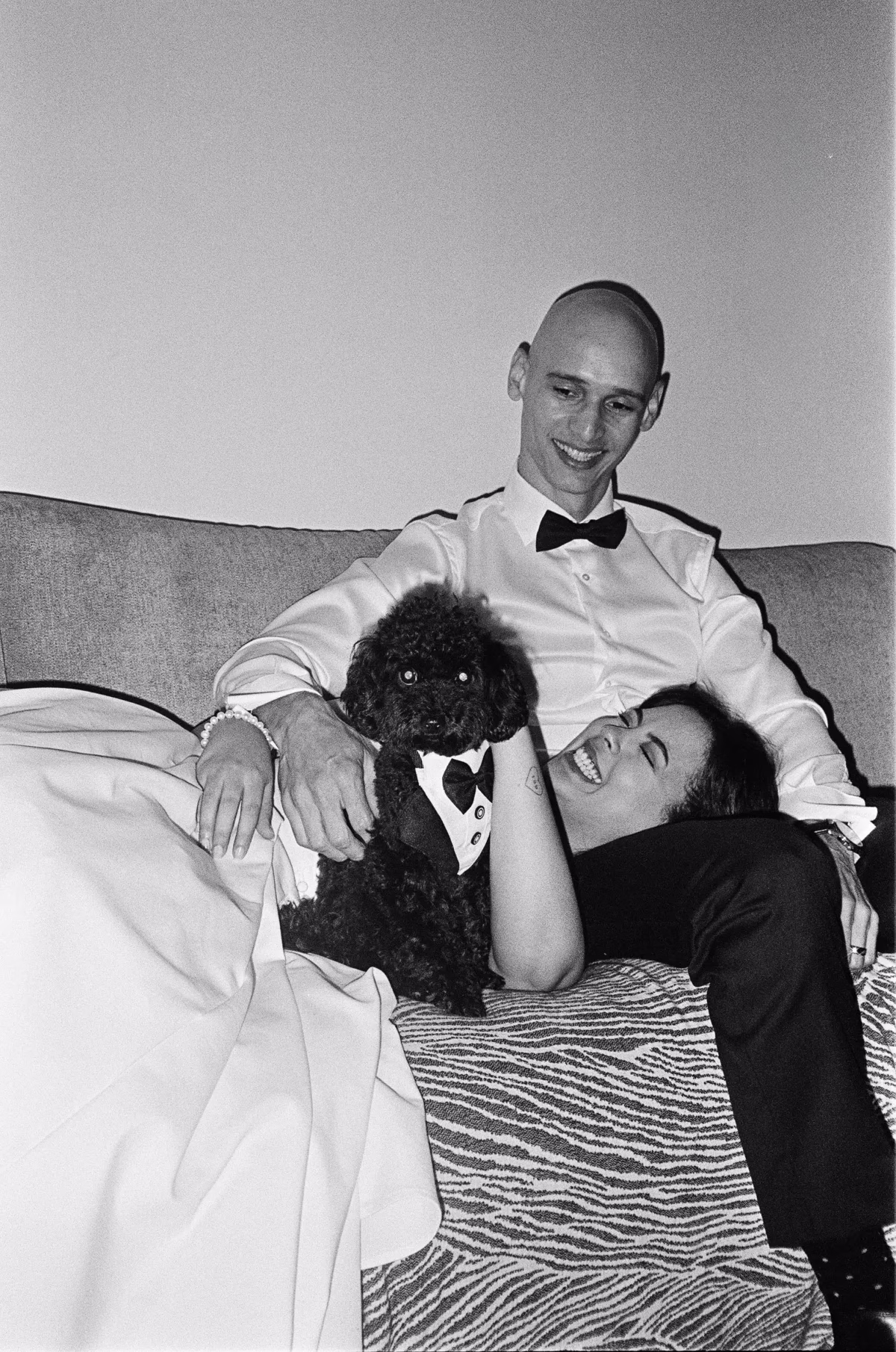 Black and white photograph of a bride reclining beside the groom with a small dog wearing a bow tie.