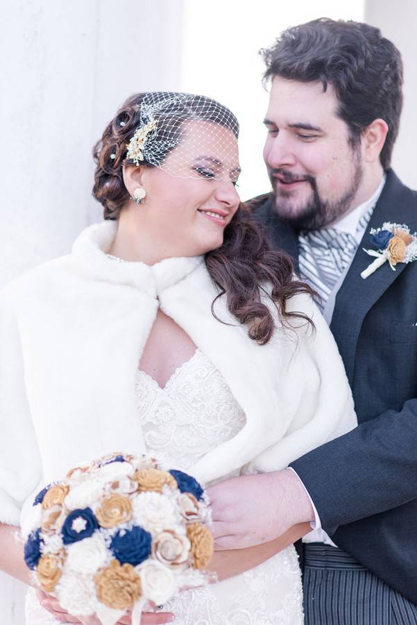 Bride in a white faux-fur capelet and birdcage veil embracing her groom, holding a sola-wood bouquet