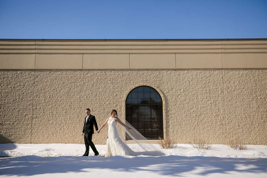 Bride in lace fit-and-flare gown with long veil trailing over fresh snow walking a sandstone wall with an arched window under a bright blue sky beside groom in black tux