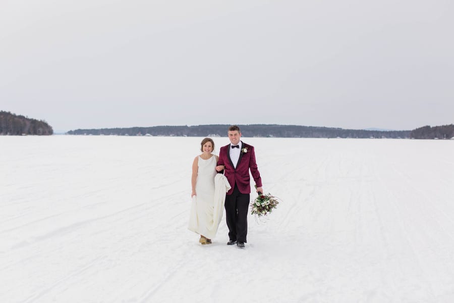 Couple walking across a vast frozen snow-covered lake bride in a sleek column gown carrying her coat groom in a burgundy velvet tuxedo jacket holding a bouquet