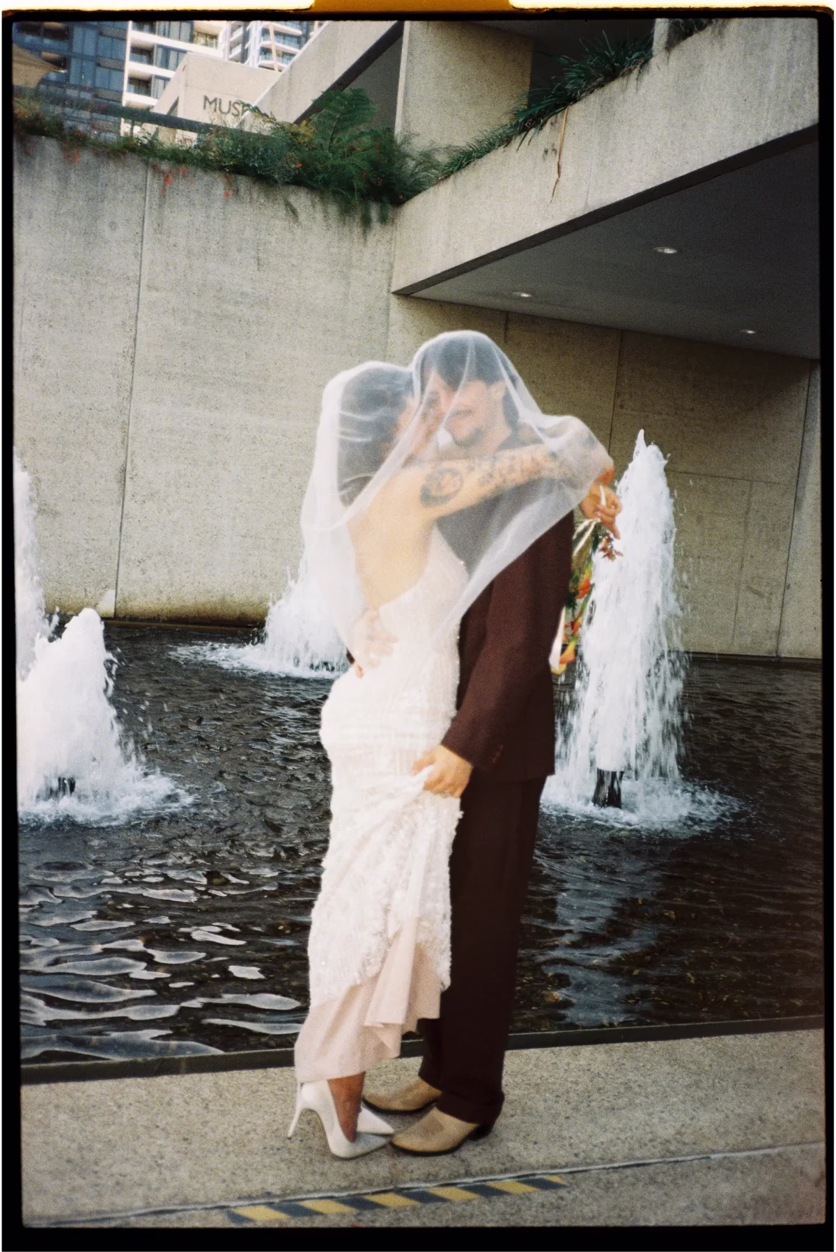 Bride and groom embracing beneath a veil beside outdoor fountains.