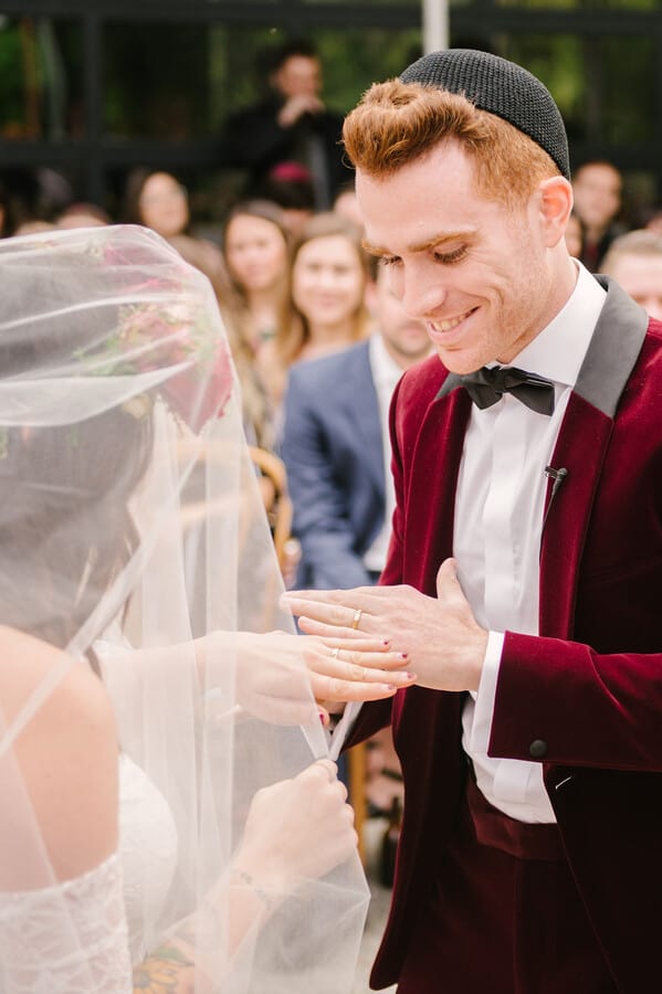 Groom in a kippah and burgundy velvet jacket smiling during the ring exchange with his veiled bride