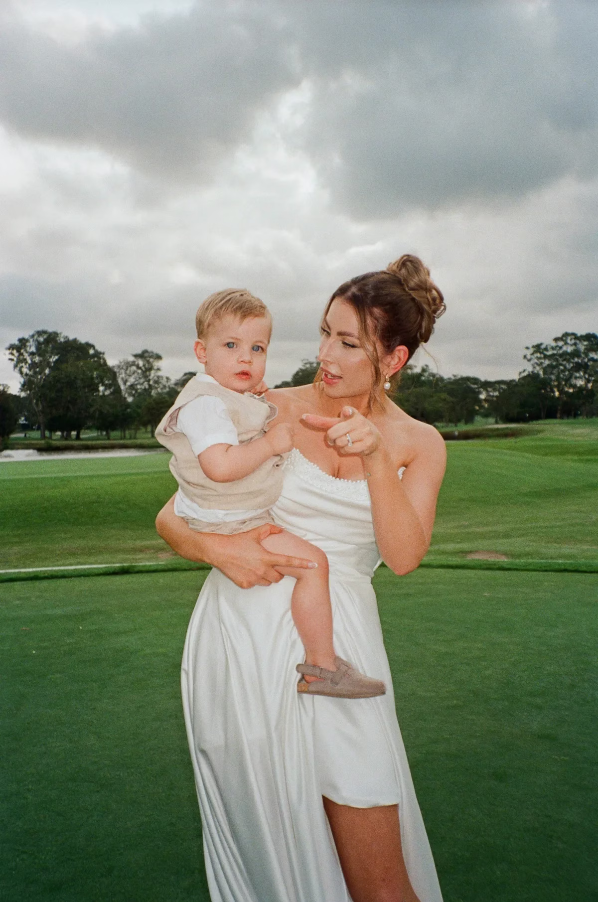Bride in a draped white dress holding a small child on a lawn beneath a cloudy sky.