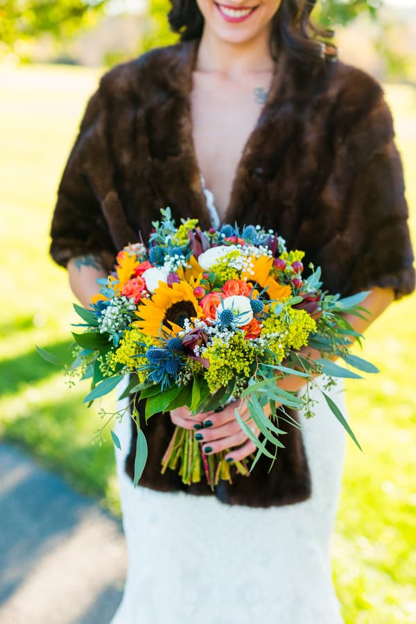 Bride in an espresso brown fur stole over a lace gown holding a sunflower and thistle bouquet in sunlight