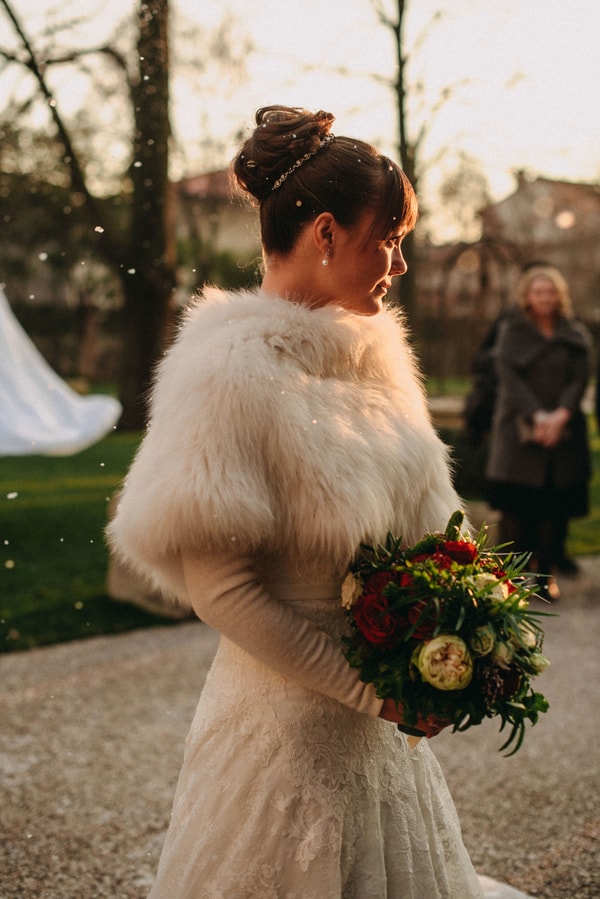 Bride in profile wearing a plush ivory fur capelet over a long-sleeve lace gown, holding a red and white bouquet