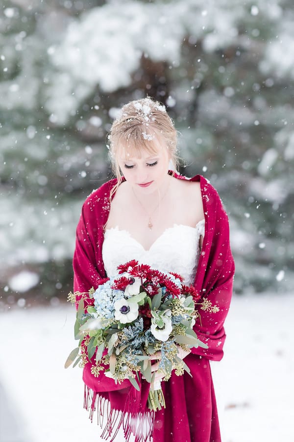 Bride in a crimson knit fringed wrap over a sweetheart lace gown, holding a red and blue bouquet as snow falls