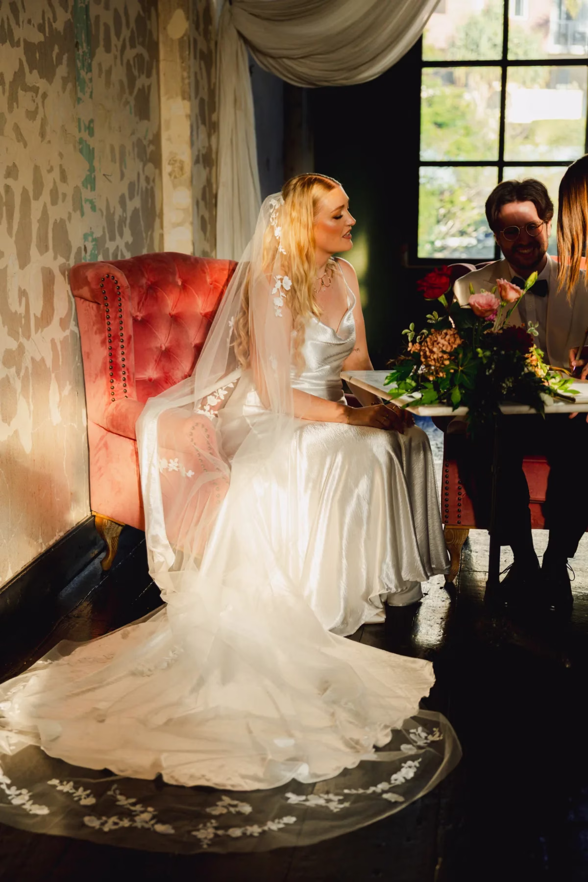 Bride seated in a red upholstered armchair beside a window, with her wedding dress spread across the floor.