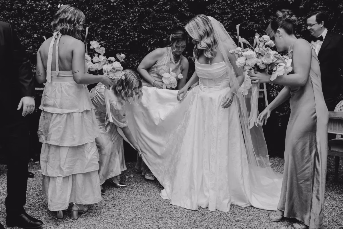 Black and white photograph of bridesmaids helping to arrange the train of a wedding dress outdoors.
