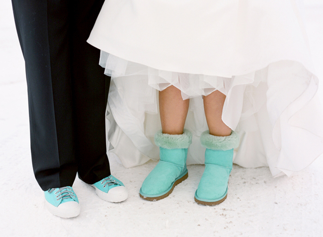 Bride's fur-topped aqua blue boots under a tulle gown next to groom's matching aqua sneakers on fresh snow