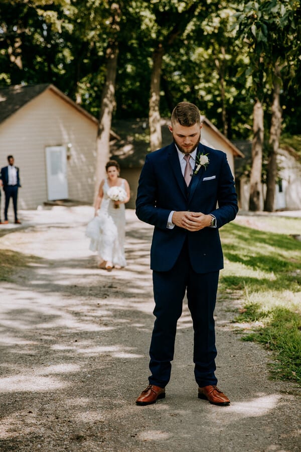 Groom in a navy suit facing away on a tree-lined path as the bride walks up behind him for a first look
