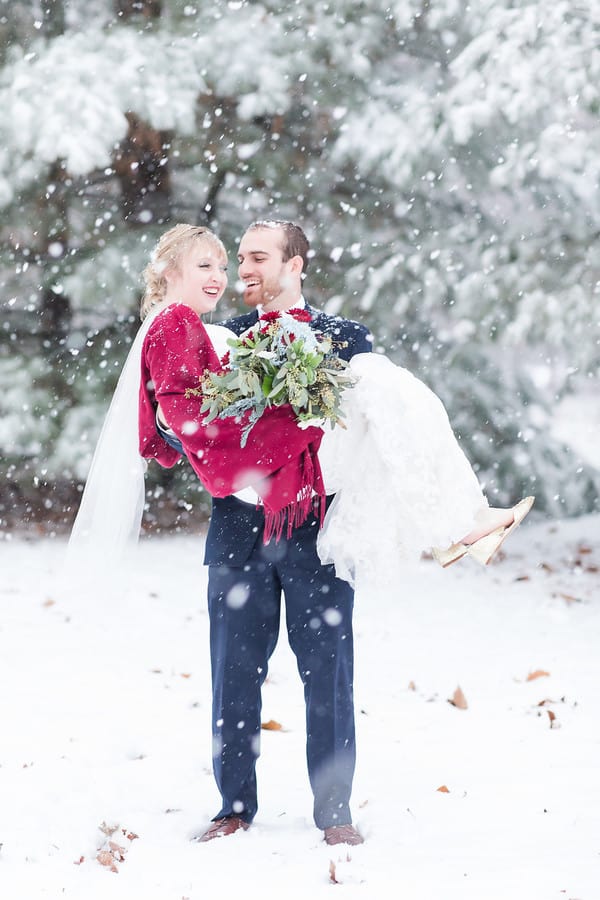 Groom in navy suit lifting laughing bride in a cranberry shawl and gold heels amid heavy falling snow with snowy evergreens behind