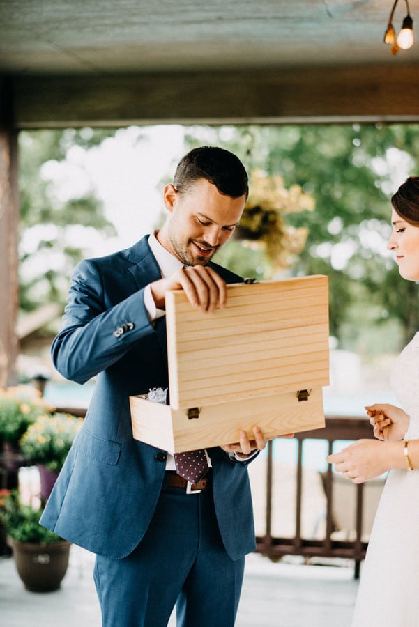 Groom in a blue suit smiling as he opens a wooden gift box while the bride watches