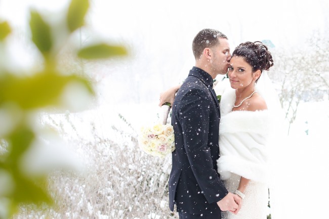 Groom kissing bride's temple as snow falls, bride in a white faux-fur shrug and pearl choker