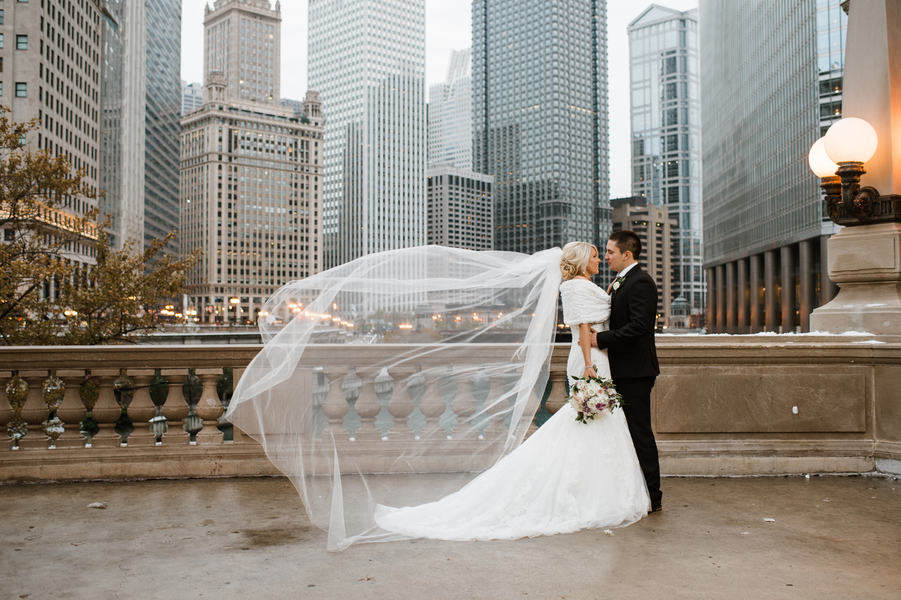 Couple on a river bridge with the Chicago skyline behind them and the bride's cathedral veil billowing across the frame bride in white fur wrap groom in black tux