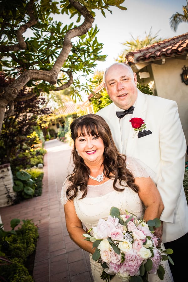 An older bride and groom standing close in a garden, the groom just behind the bride who holds a soft pink bouquet