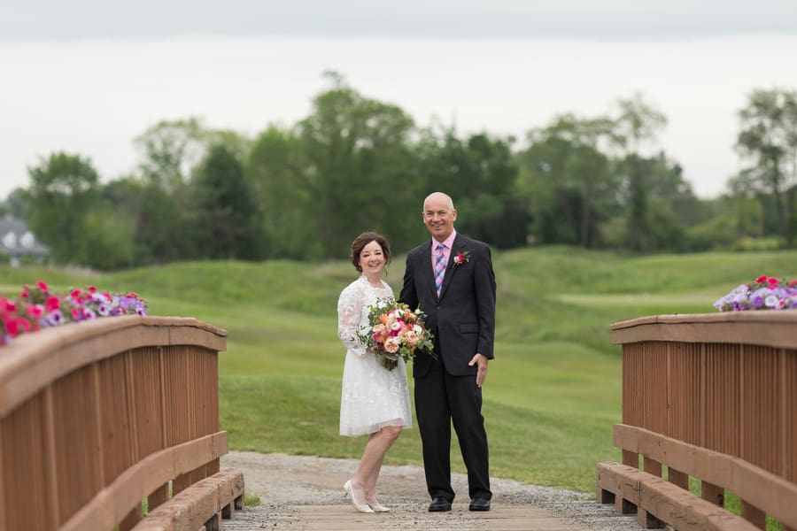 An older bride and groom posed full length on a flower-lined wooden bridge, the groom's arm around the bride