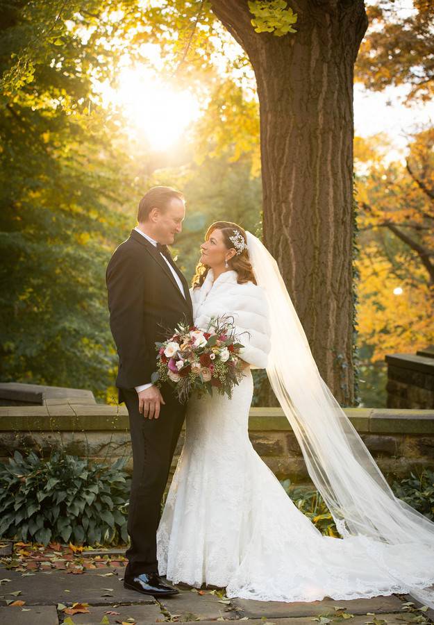 Bride in lace mermaid gown with ivory fur shrug and long cathedral veil holding a mauve and burgundy bouquet beside groom in black tux in golden light