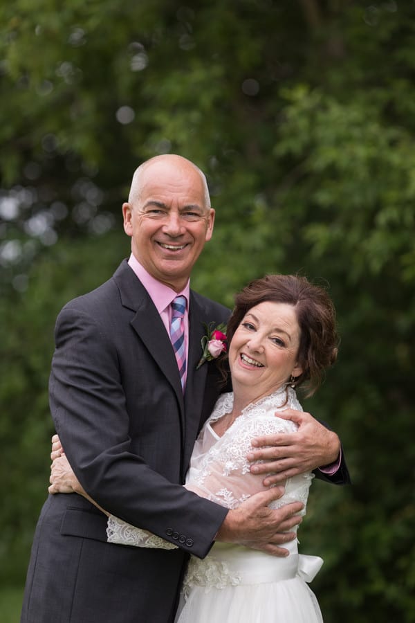 An older bride and groom in a close embrace, arms around each other, smiling at the camera against green trees