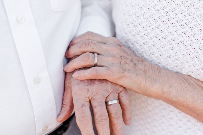 Close-up of an elderly couple's clasped hands resting together, both wearing worn wedding bands