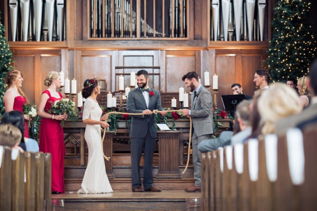 Bride in long-sleeve lace gown and groom in gray suit at candlelit altar with two lit Christmas trees and crimson bridesmaids