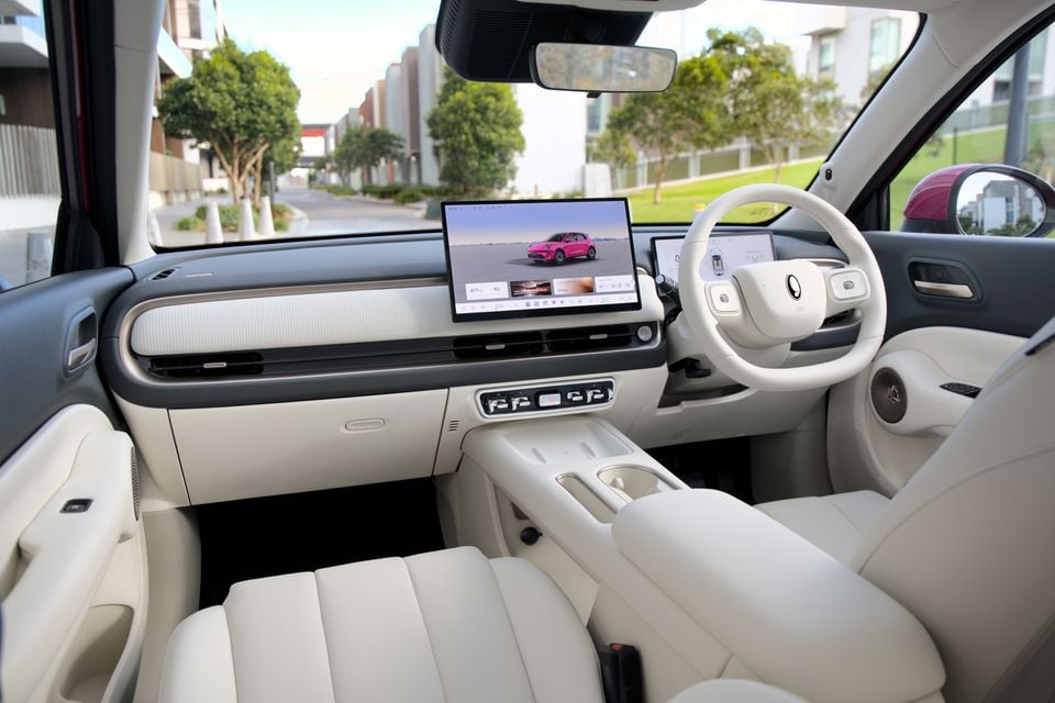 Interior of the 2026 GWM ORA 5 showing the white cabin, large central touchscreen displaying a pink car, flat-bottomed steering wheel, and front seats viewed from the passenger side