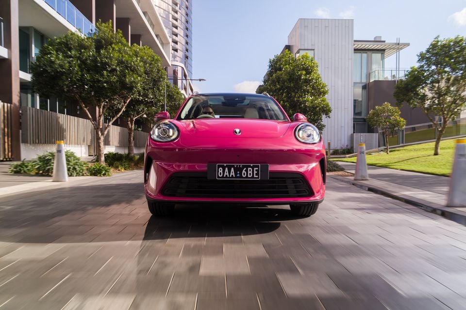 A pink GWM ORA 5 small SUV seen from the front, driving through an urban street in Australia