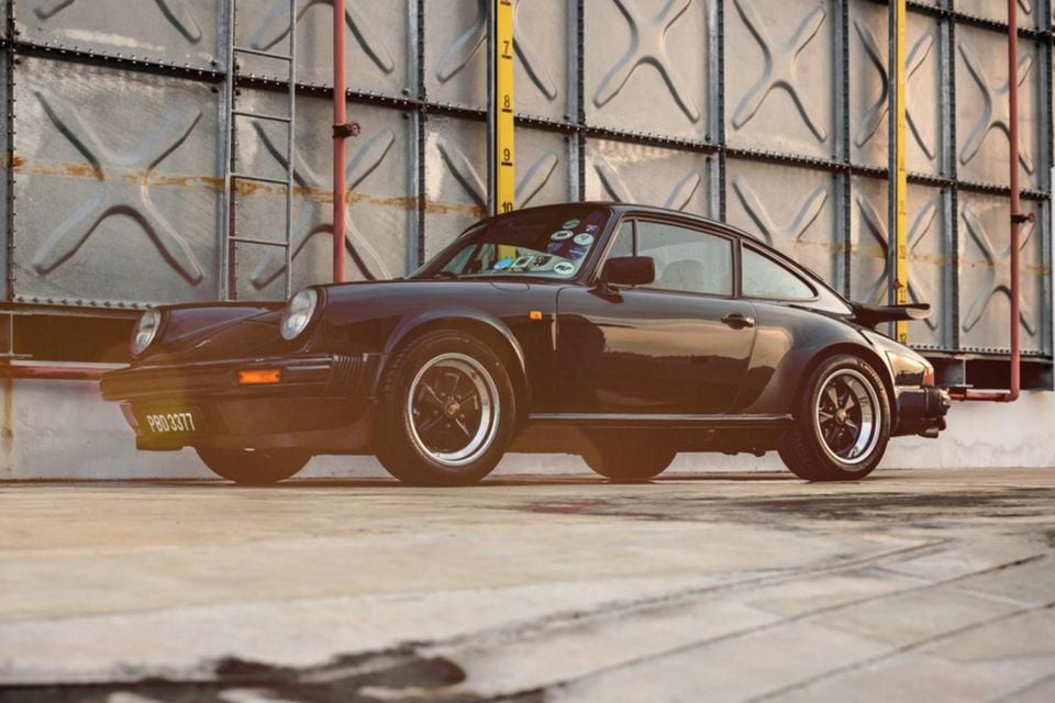 A black 1981 Porsche 911 SC coupe parked in front of a riveted metal water tank, seen from the front left side