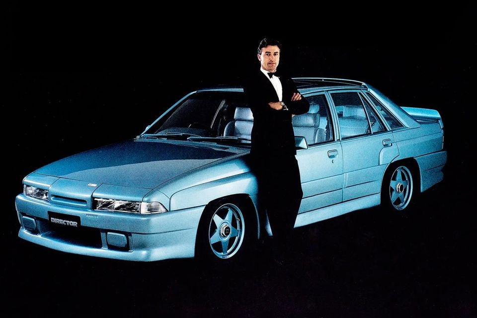 A light blue 1987 HDT VL Holden Calais Director sedan seen from the front, with Peter Brock in a tuxedo leaning against it