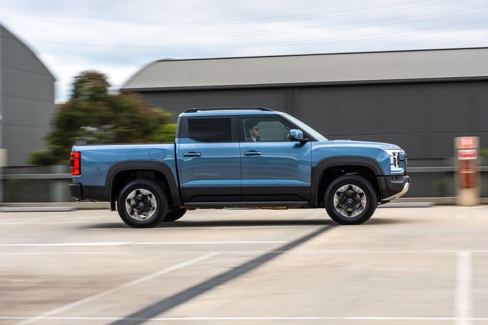 A blue dual-cab ute driving through a car park, seen side-on in profile with a motion-blurred background.