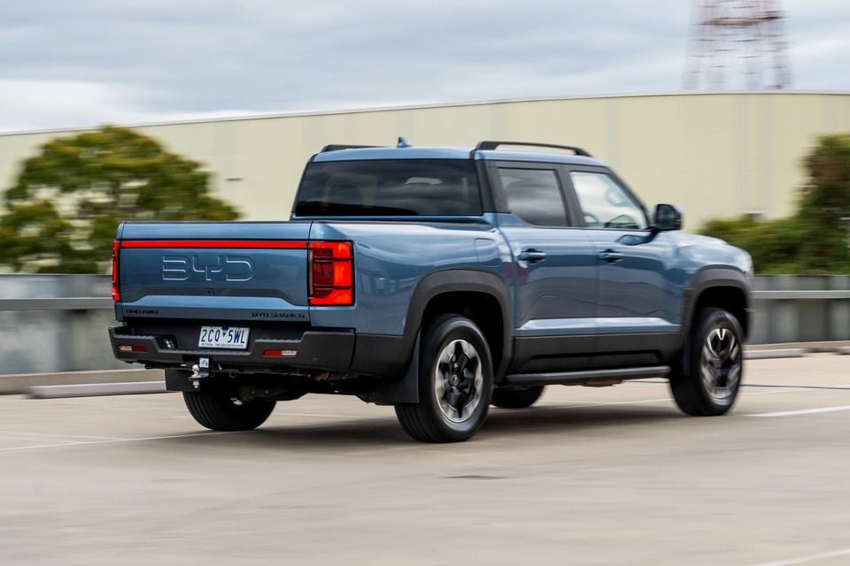A blue-grey 2026 BYD Shark 6 dual-cab ute driving across a rooftop car park, seen from the rear passenger side